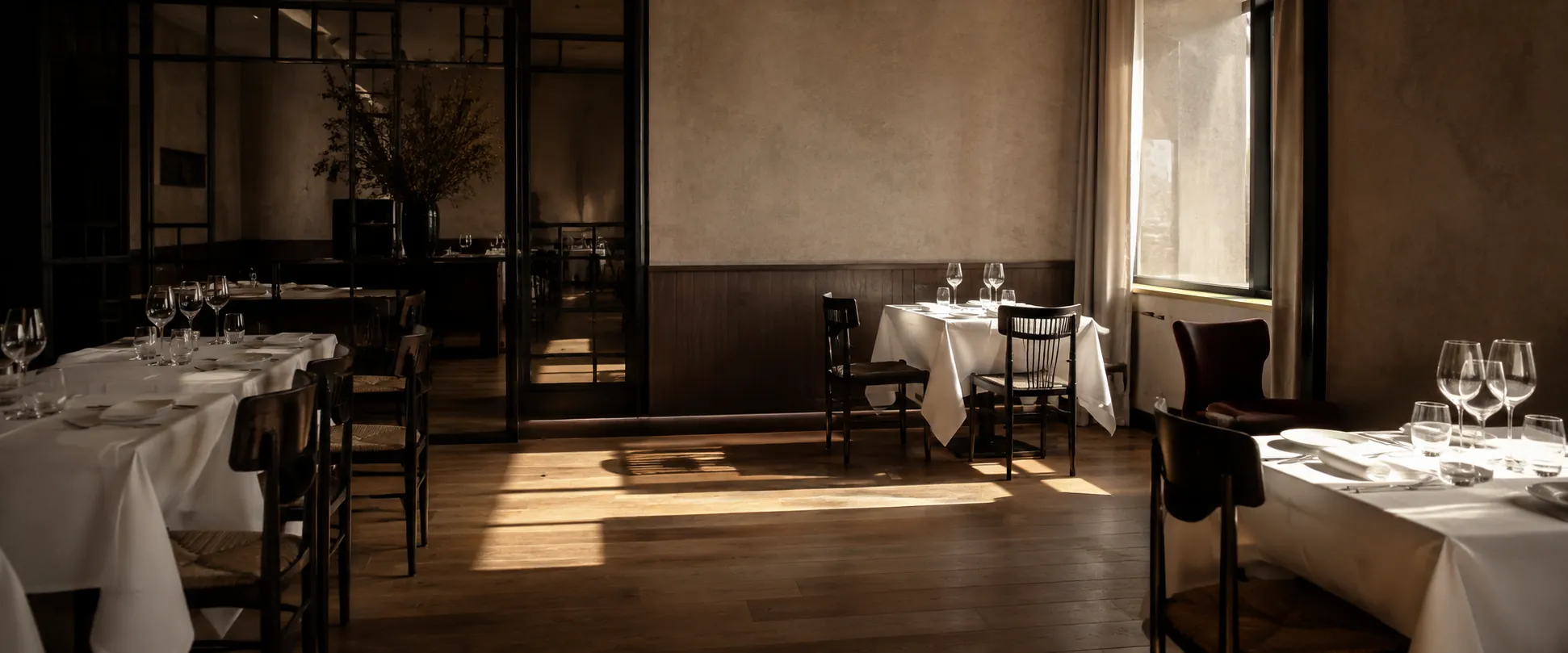 An empty upscale dining room stands ready for service as late-afternoon light stretches across the dark wooden floor.